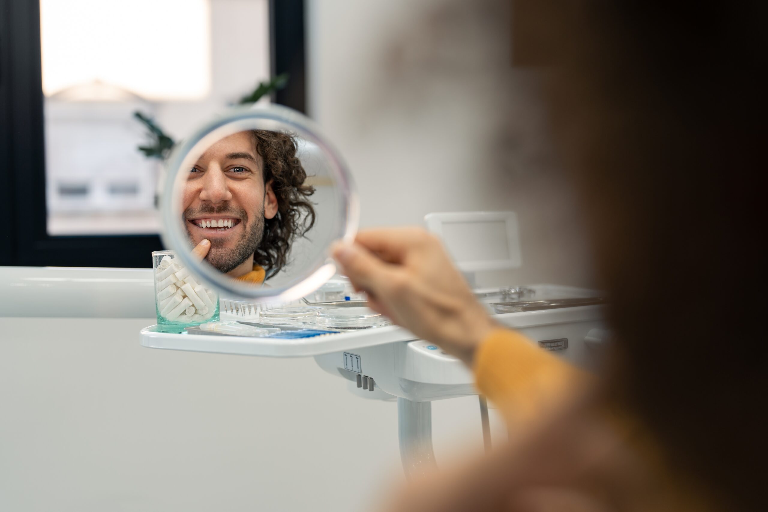 Handsome young man looking in mirror at dentist checking results after cosmetic dental treatment, feeling happy and confident